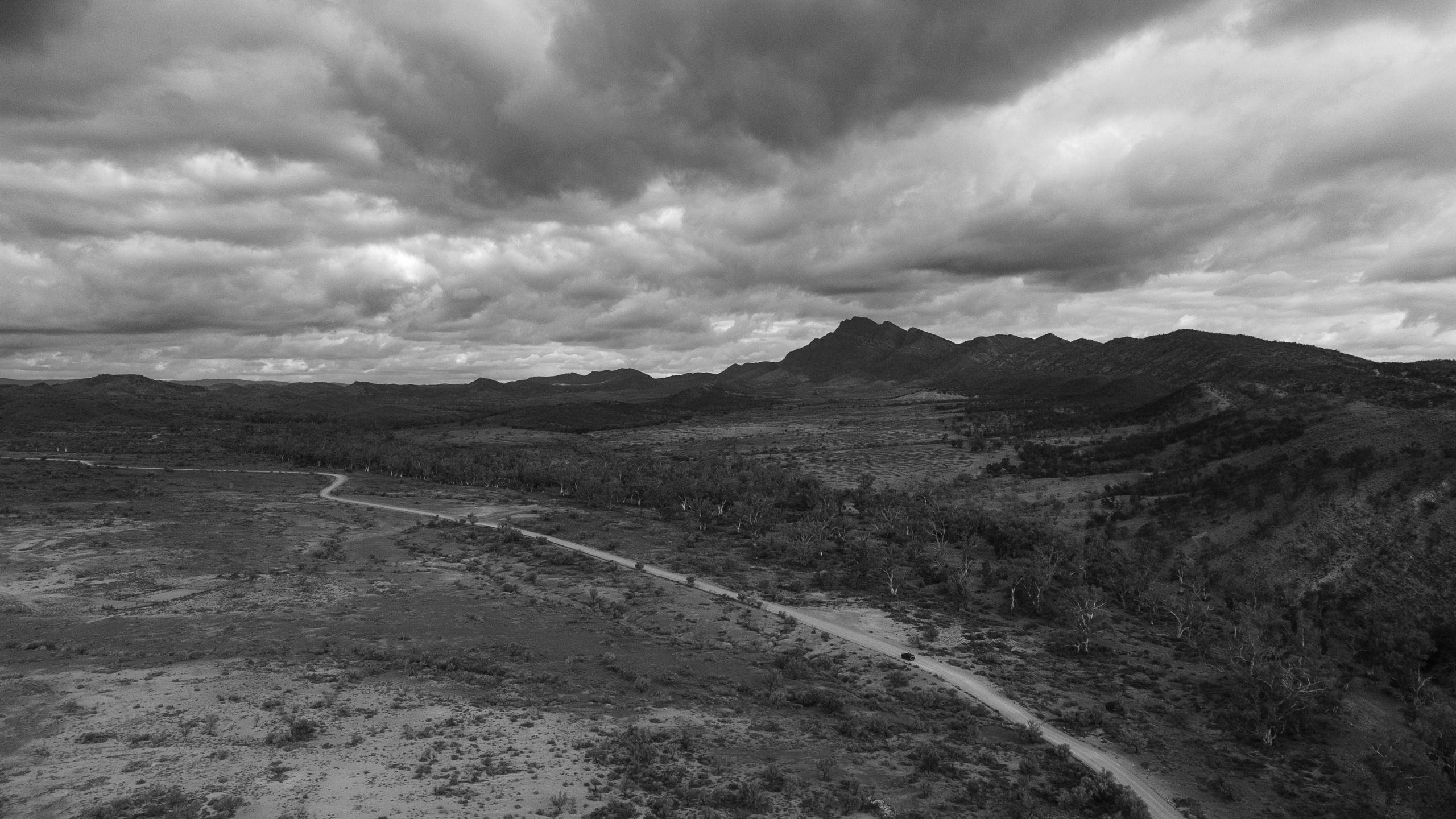 Black and white image capturing a four-wheel drive on the Moralana Scenic Drive, framed by dramatic clouds and the silhouette of the Flinders Ranges.
