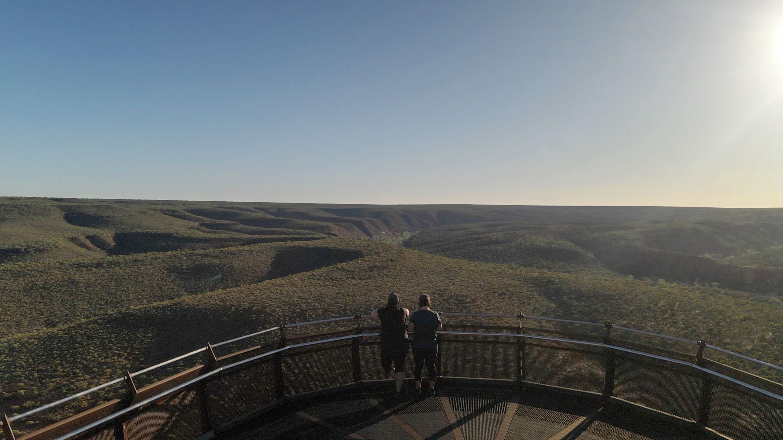 Two women standing on the Kalbarri Skywalk in Kalbarri National Park, Western Australia, overlooking the Murchison River Gorge as the morning sun casts a warm glow.