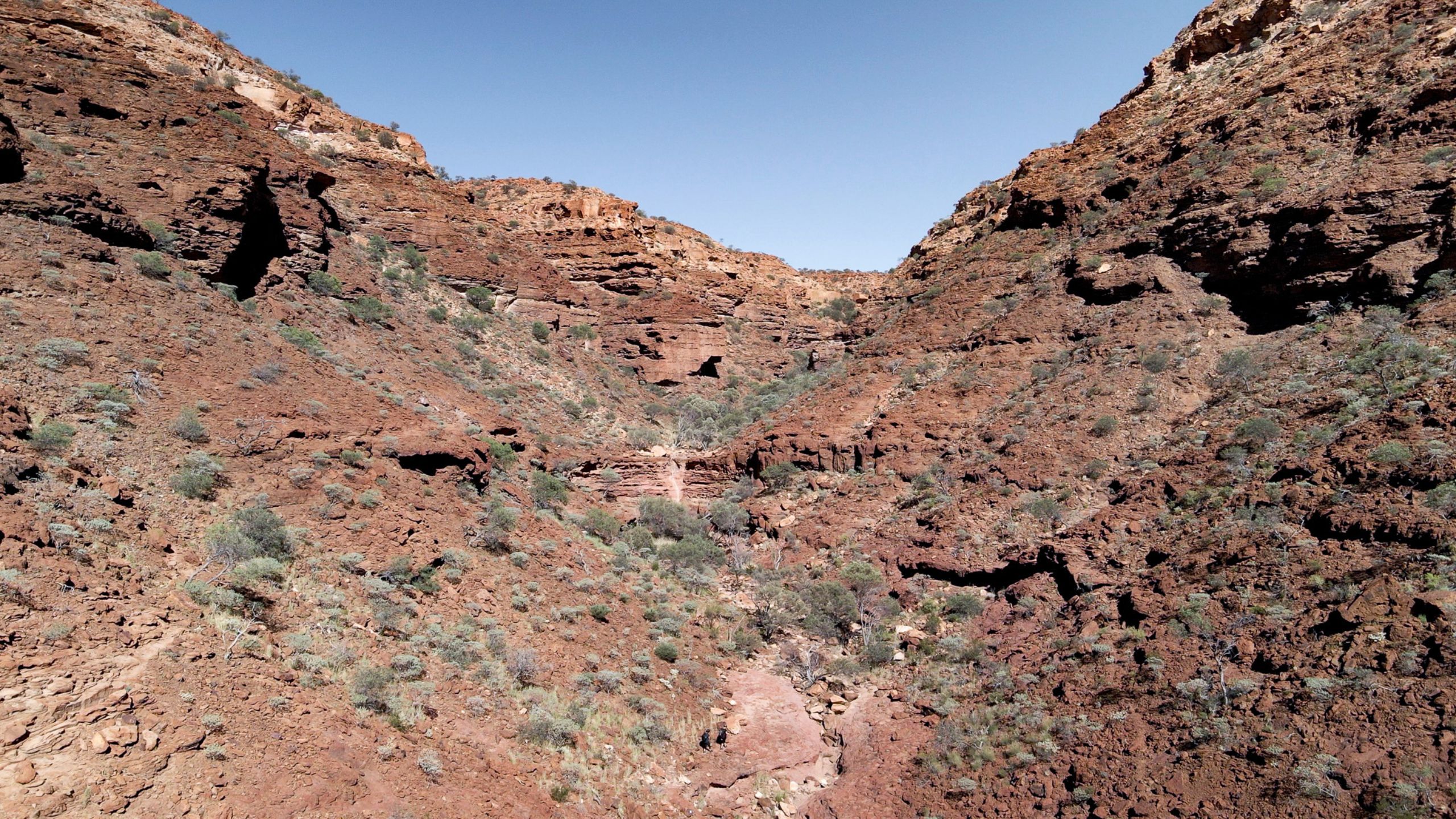 Two hikers walk along a rugged trail beneath the towering, red sandstone cliffs of Drapers Gorge in Kennedy Range National Park, Western Australia, under a bright blue sky