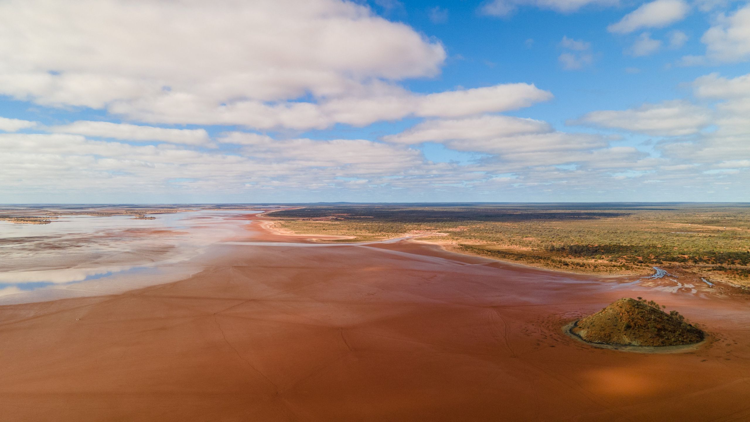 Aerial view of Lake Ballard with shallow water covering parts of the salt flats, reflecting the clouds and blue sky above.