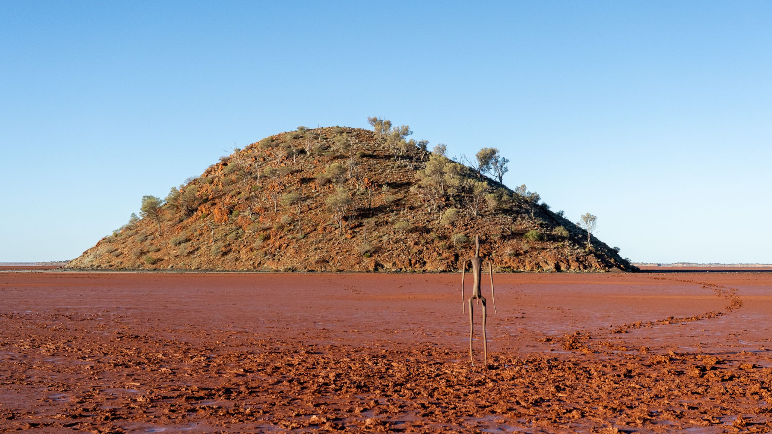 Sculpture from Antony Gormley’s "Inside Australia" installation stands in the foreground of the salt lake at Lake Ballard, with one of the Seven Sisters Hill rising in the distance.