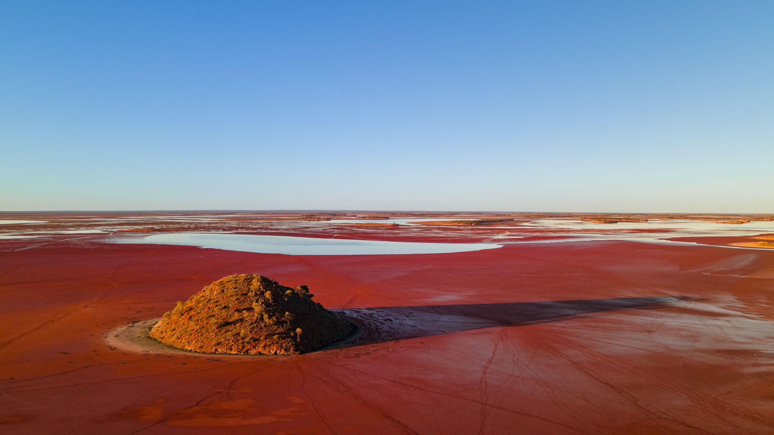 Aerial view of Lake Ballard at sunset, with deep red mud in the foreground, low water visible in the distance on the salt flats, a vivid blue sky above, and long shadows stretching across the landscape